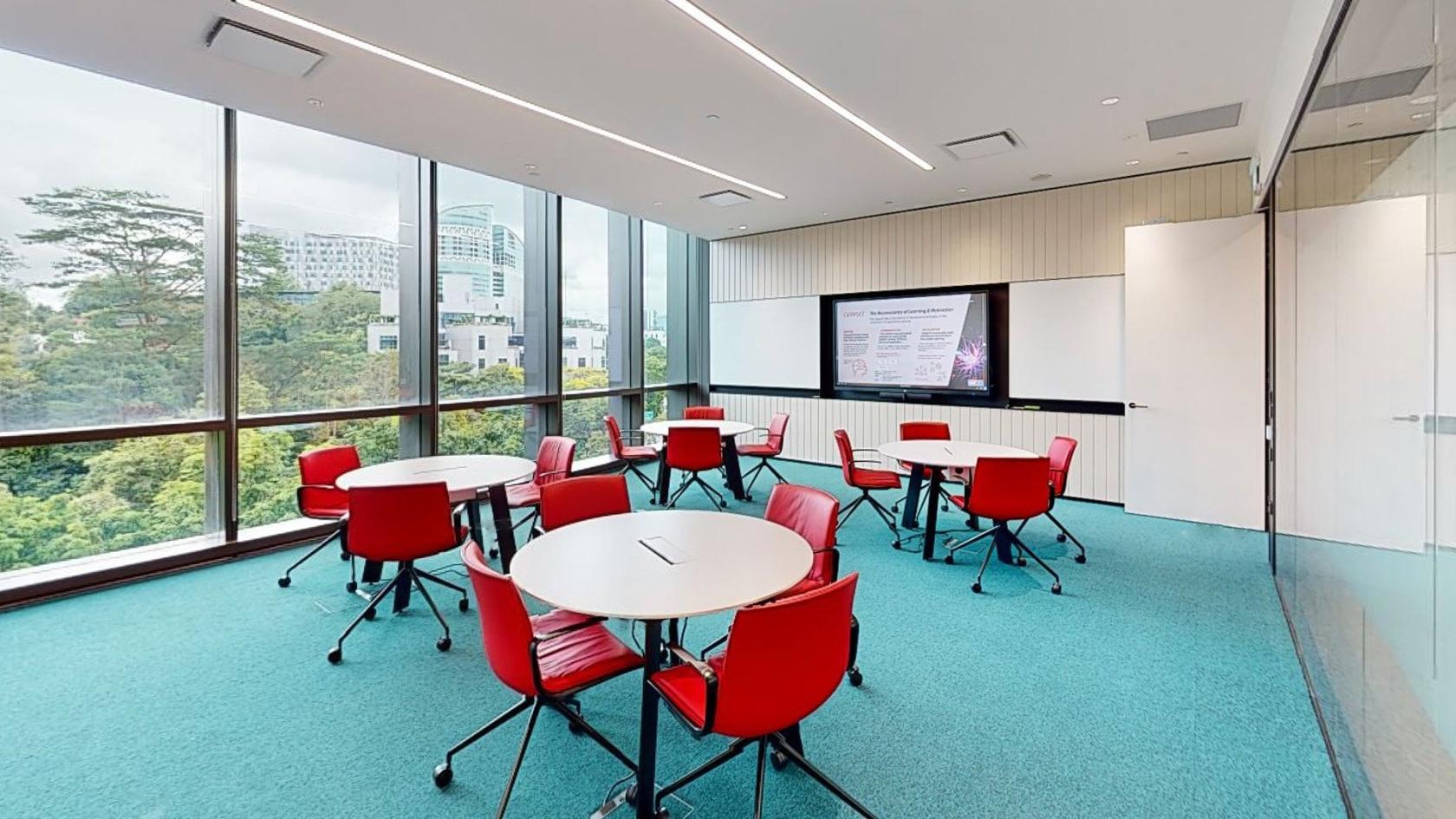 A conference room with floor-to-ceiling windows, round tables, red chairs, and a wall-mounted screen at Catapult.