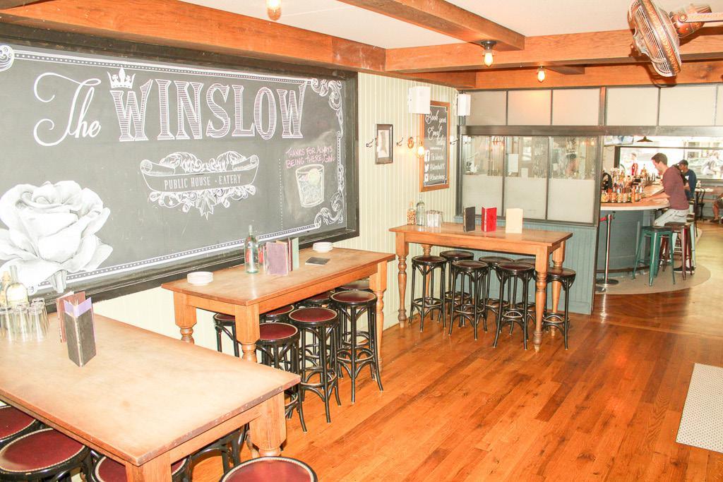 A bar with communal high-top tables, wooden floors, stools, and a chalkboard wall at The Winslow.