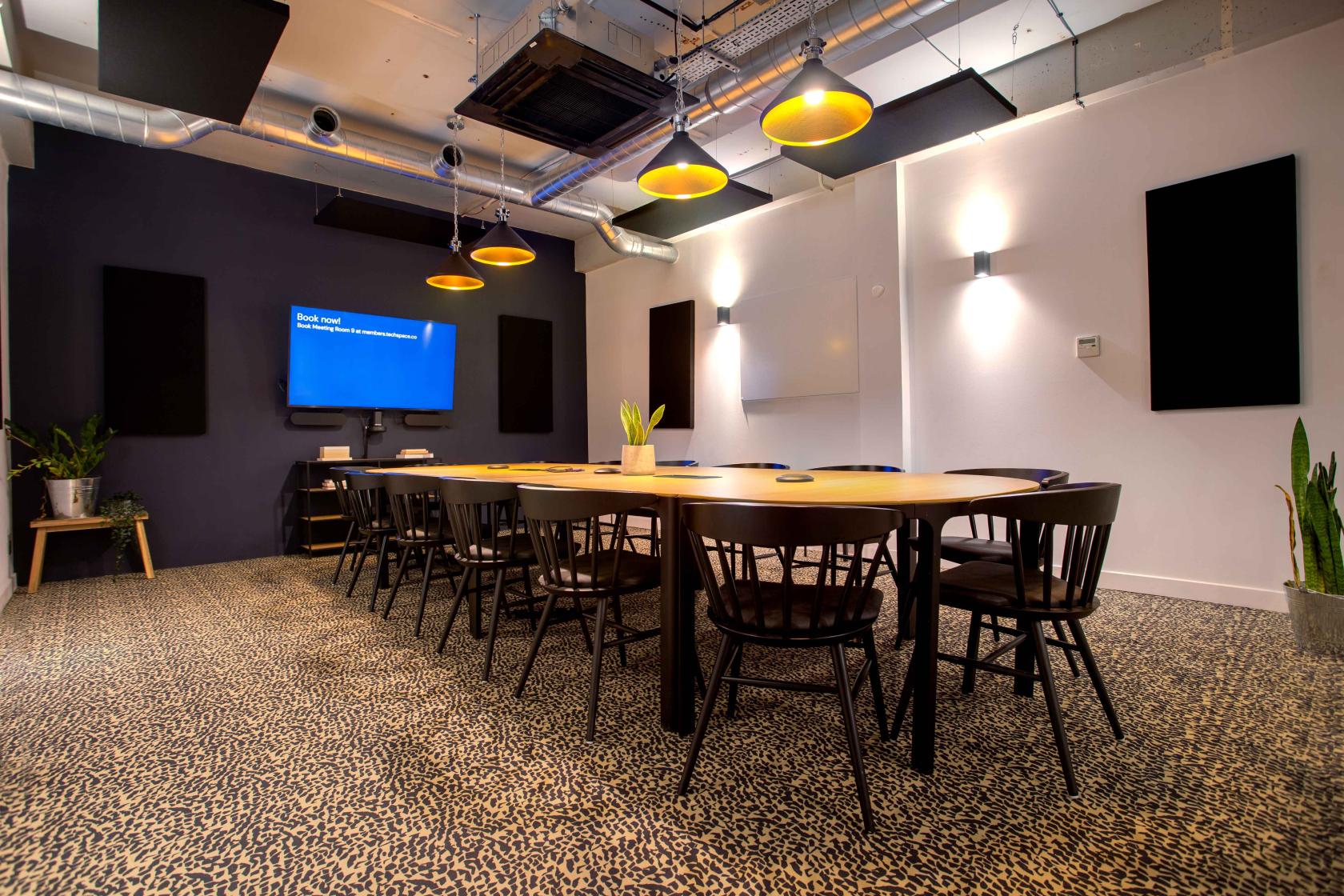 A meeting room with an oval conference table, black chairs, pendant lights, and wall-mounted screens at Techspace Shoreditch.