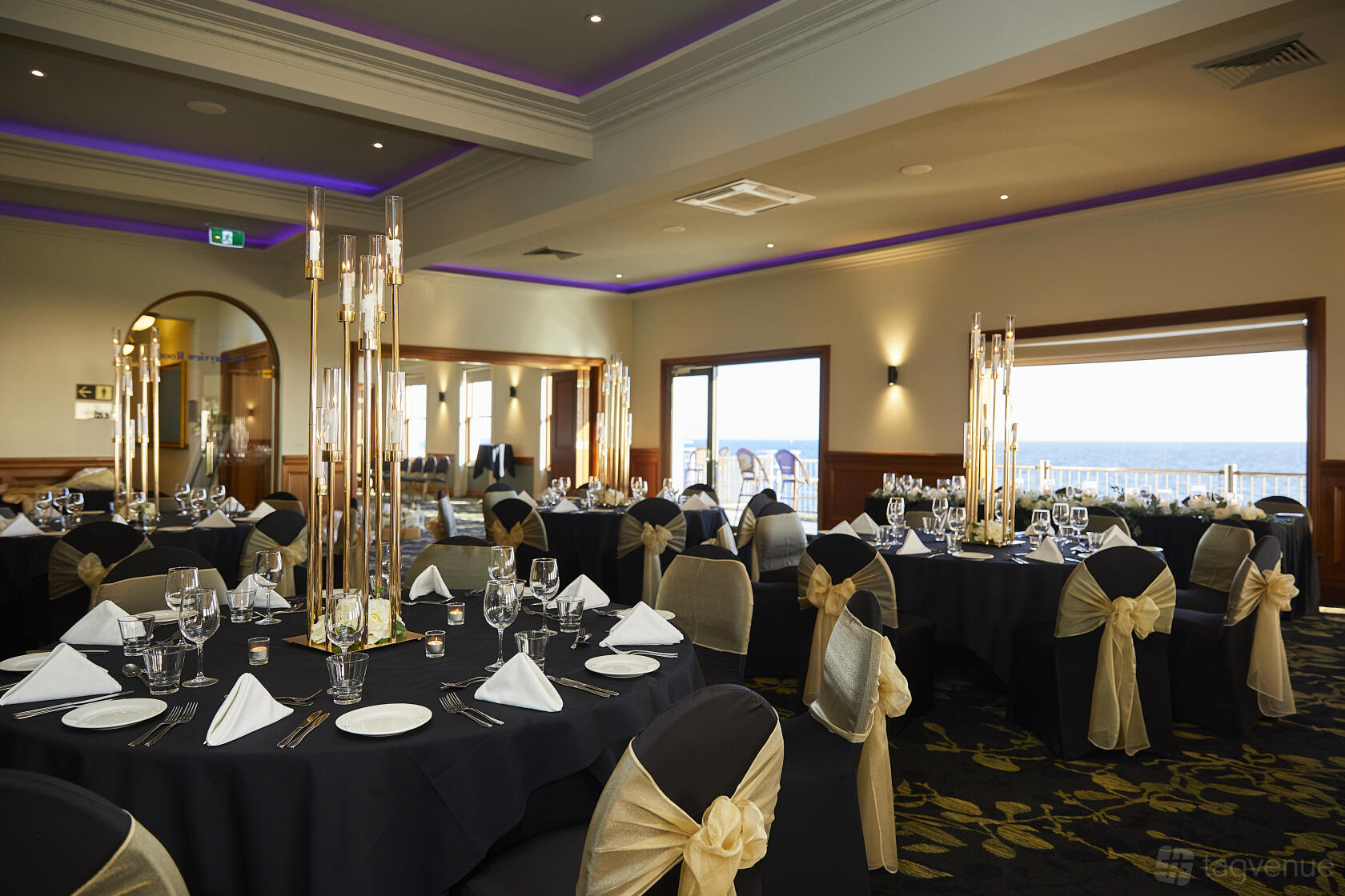 A pub event space with round tables dressed in black linens and gold sashes, large windows overlooking the ocean at Brighton Beach Hotel.