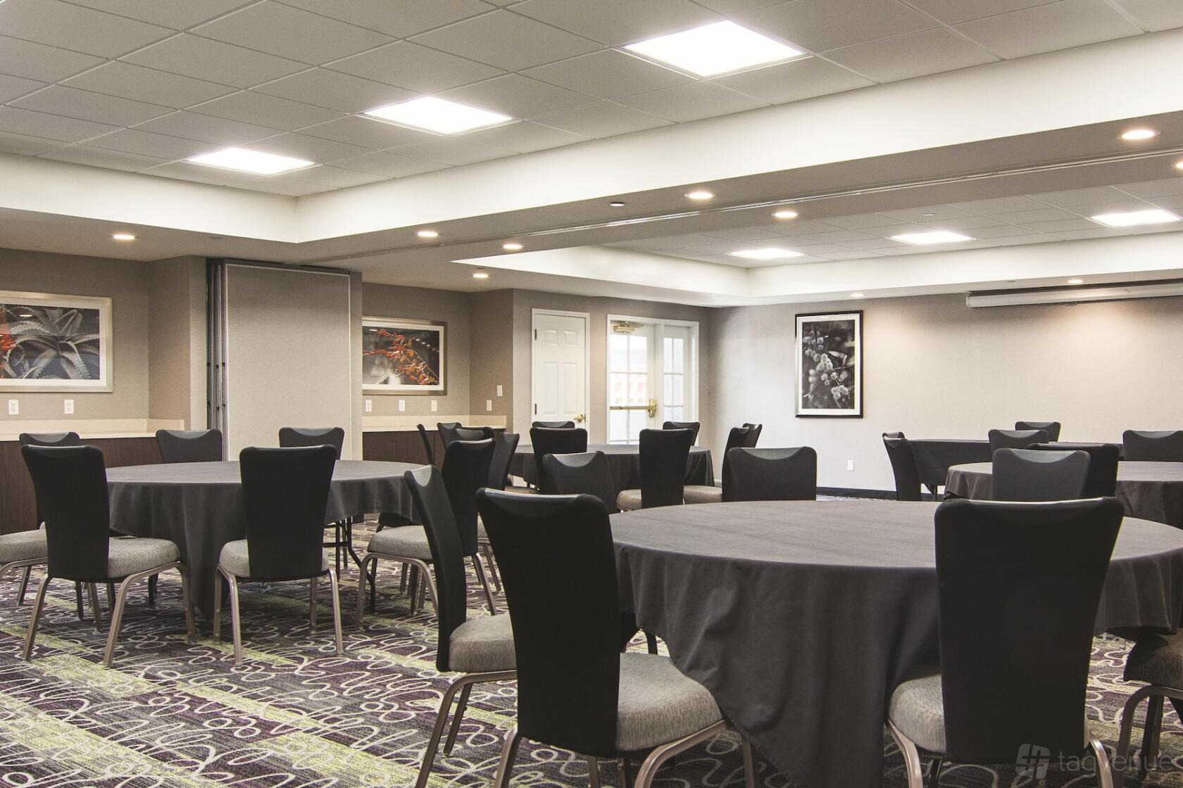 A meeting room with round tables, black chairs, carpeted floors, and framed artwork at La Quinta Inn & Suites by Atlanta Perimeter.