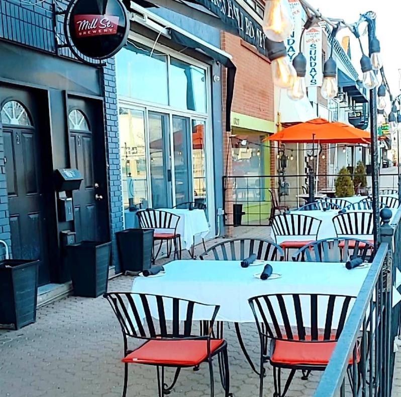 An outdoor patio with striped chairs, white tablecloths, and orange umbrellas at Clarkson Bistro Mediterranean Cuisine.