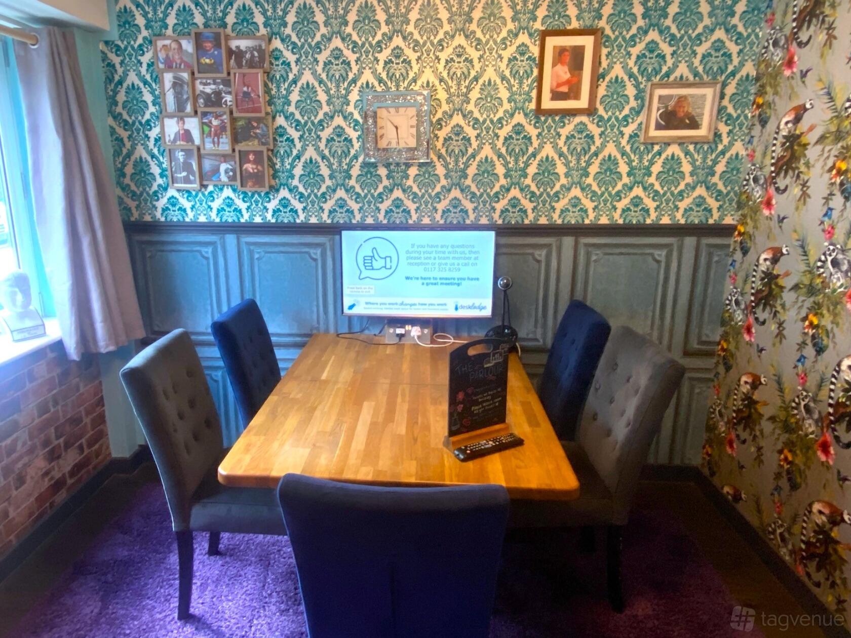 A meeting room with patterned wallpaper, a wooden table, velvet chairs, and a wall-mounted screen at DeskLodge House Bristol.