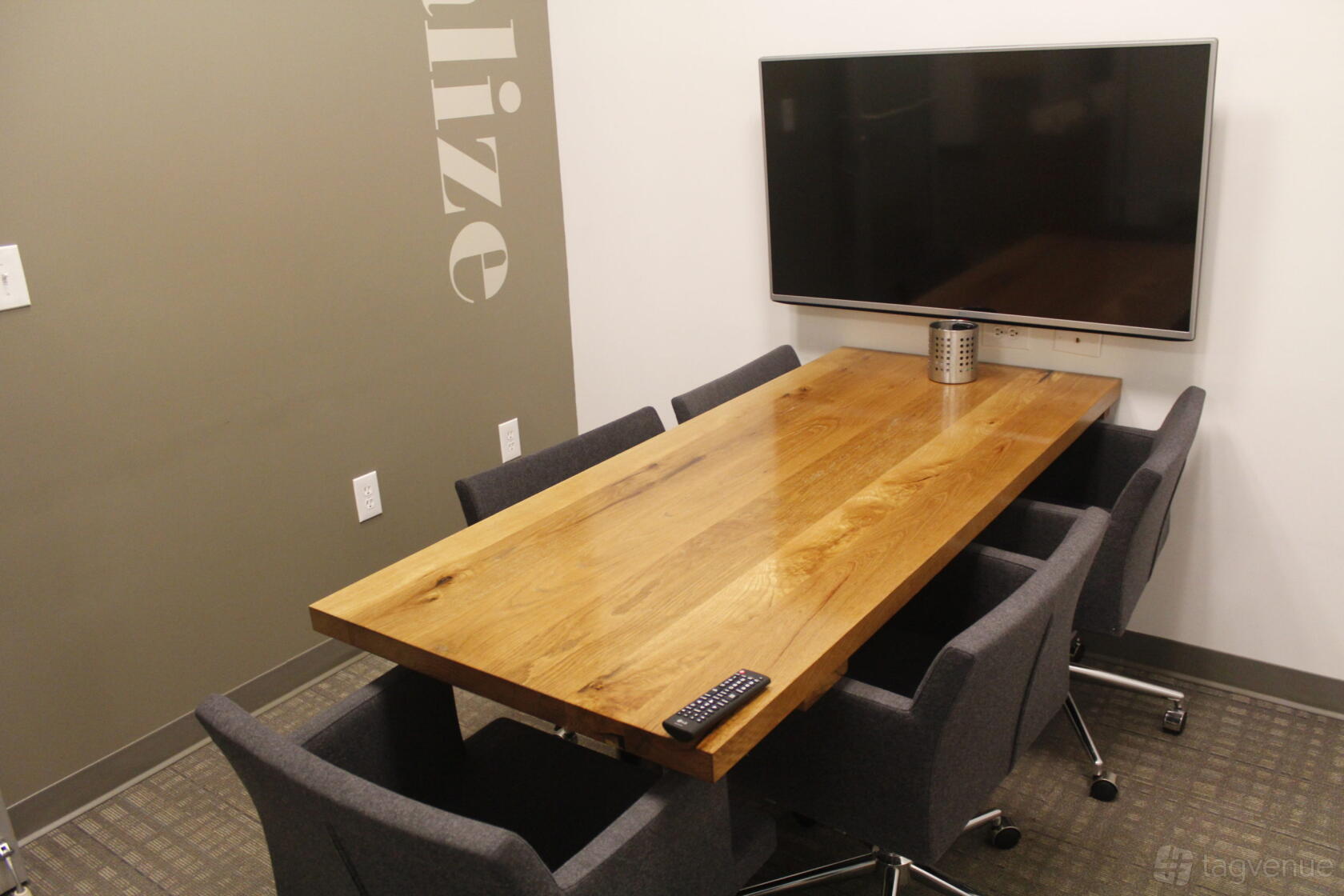 A meeting room with a wooden table, four gray chairs, wall-mounted TV, and neutral decor at ROAM ALPHARETTA.