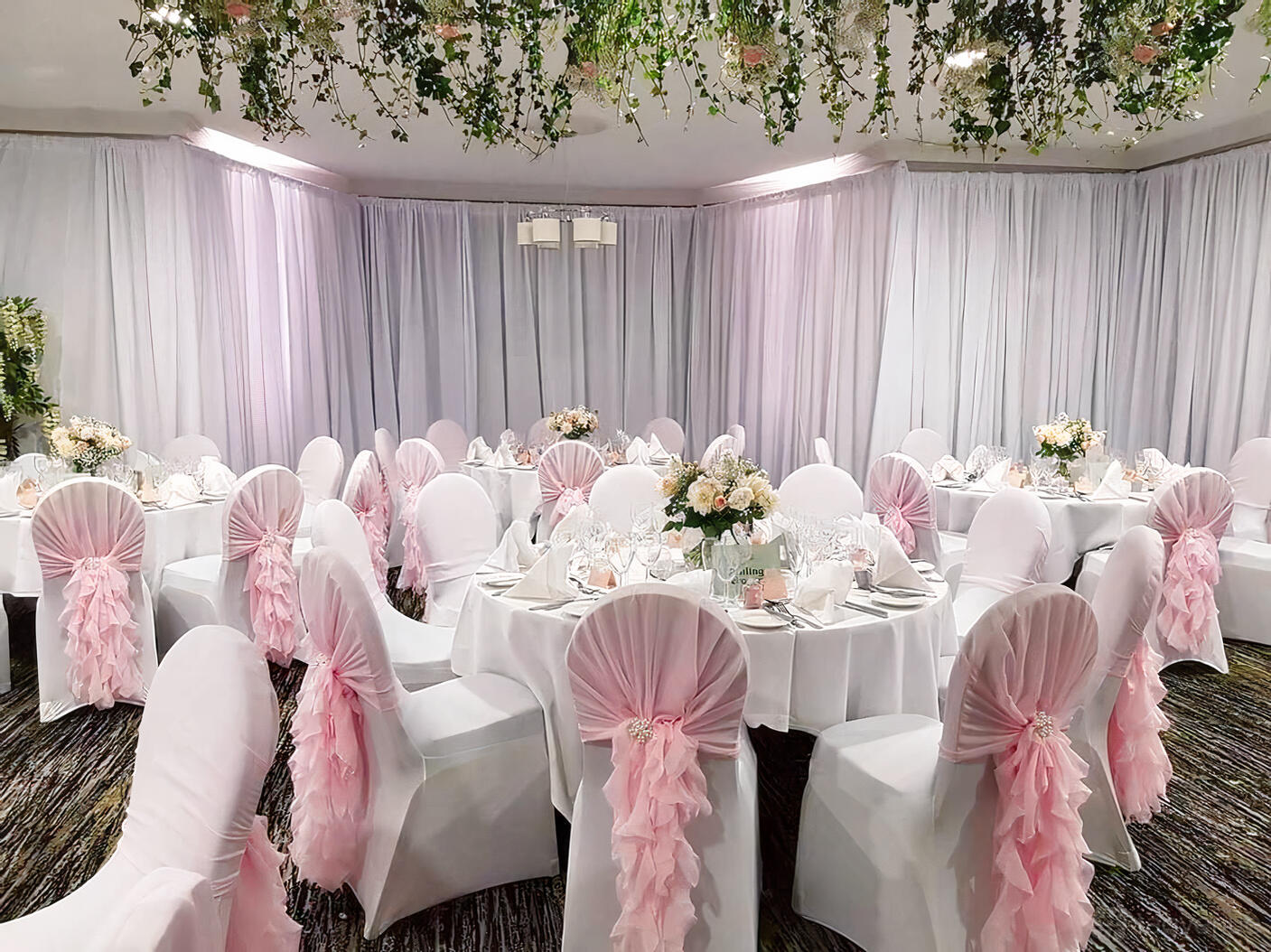An event space with round tables, white linens, pink chair sashes, floral centerpieces, and hanging greenery at Dunston Hall.