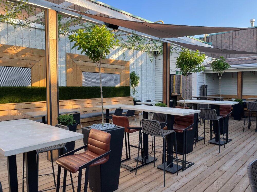 An outdoor restaurant patio with wooden decking, shade sails, potted trees, and bar-height tables at Rogue Event Space.
