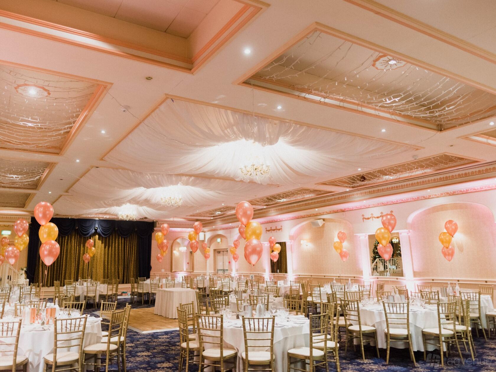 A ballroom with gold chiavari chairs, round tables, pink balloon centerpieces, and draped ceilings at New Continental Hotel.