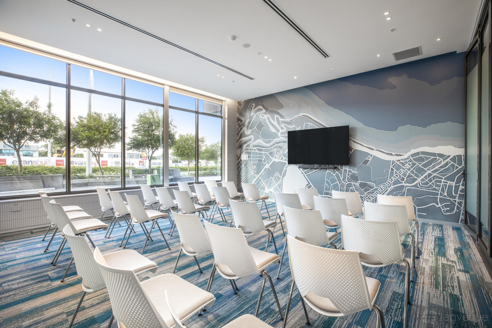 A meeting room with rows of white chairs, floor-to-ceiling windows, and a wall-mounted screen at Holiday Inn Express & Suites Sunshine Coast.