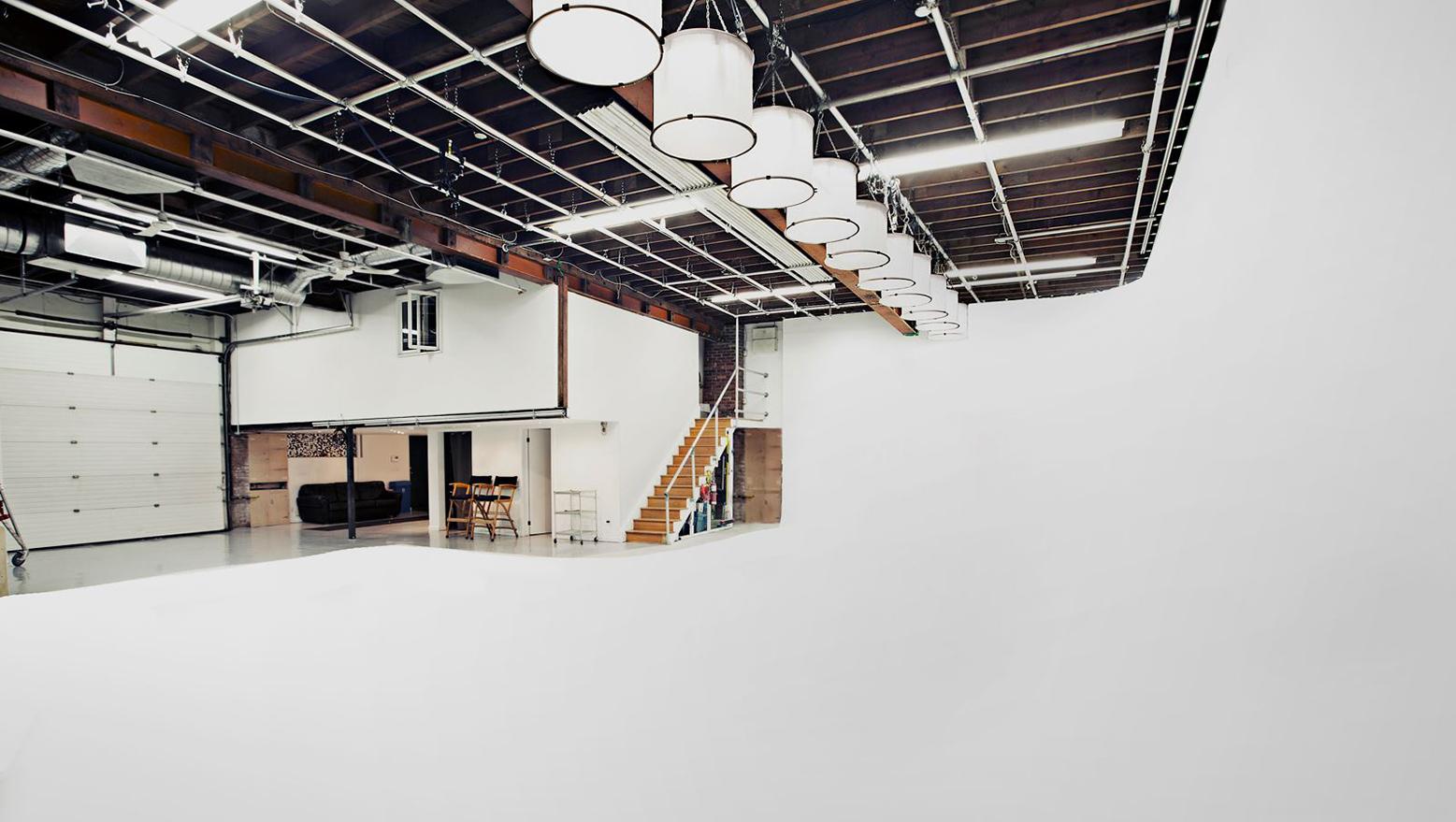 A photo and film studio with a large white cyclorama wall, exposed ceiling beams, and hanging drum lights at 276 Sterling Studio.