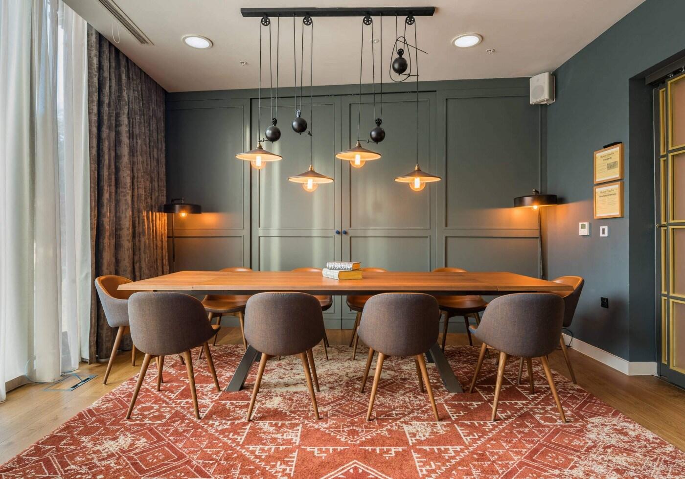 A dining room with a long wooden table, upholstered chairs, pendant lighting, and patterned rug at Hotel Indigo Manchester - Victoria Station.