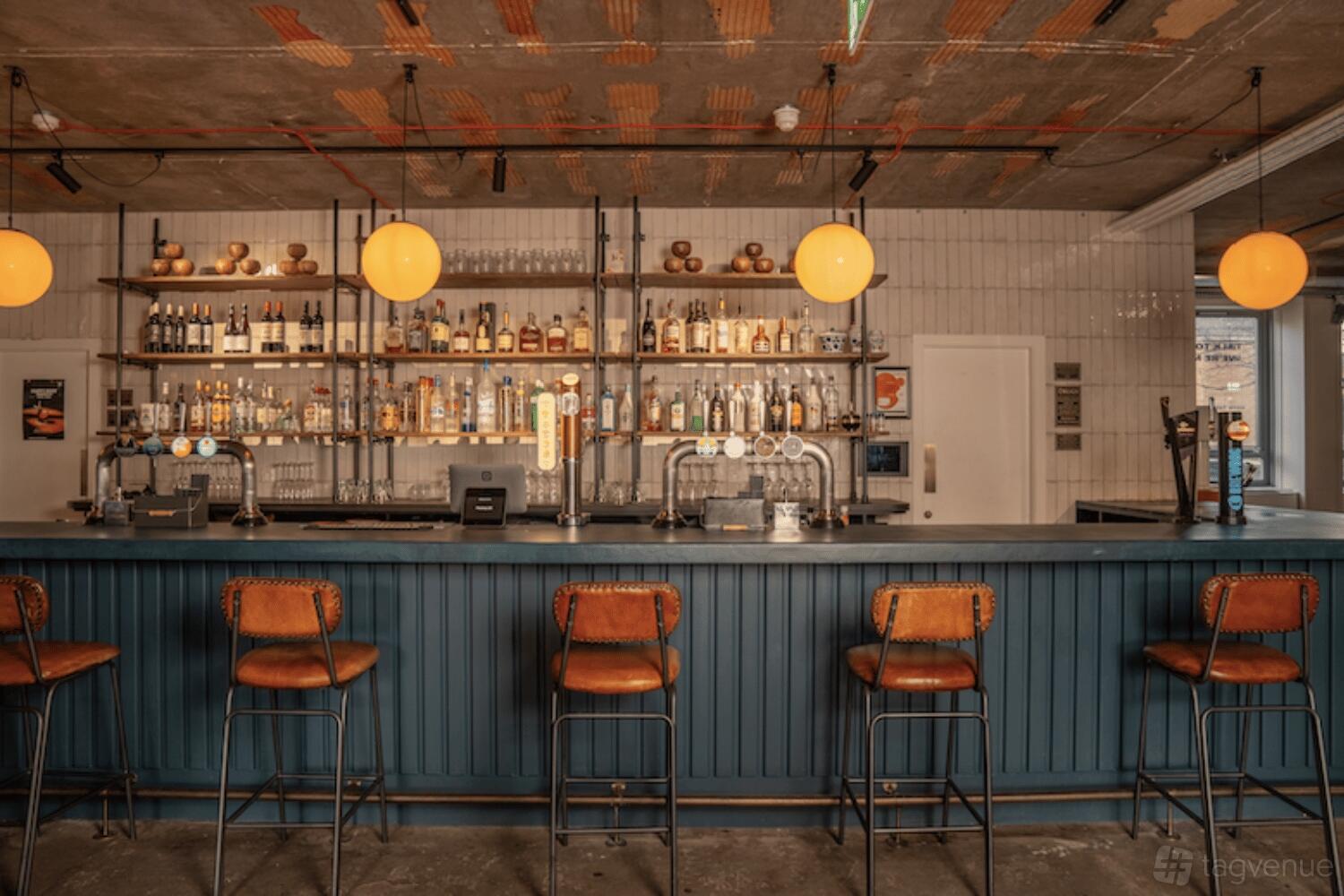 An event space with a blue paneled bar, leather barstools, globe pendant lights, and open shelving at MAKE IT Barking.