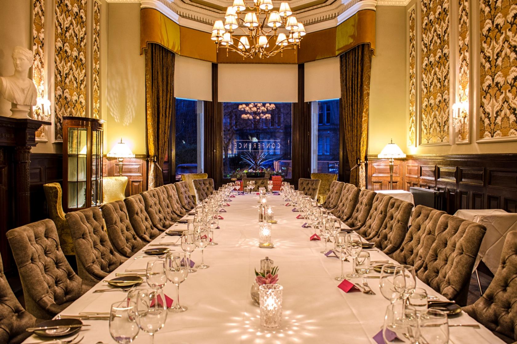 A hotel dining room with ornate wallpaper, a long banquet table, chandeliers, and tufted chairs at The Bonham Hotel.