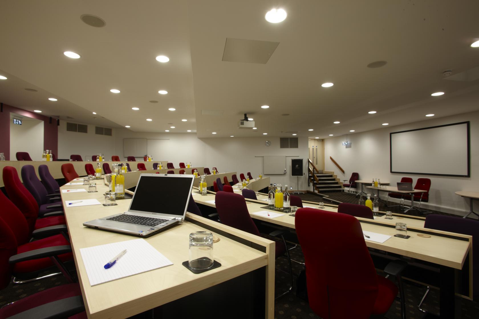 A tiered meeting room with red chairs, individual desks, and a large presentation screen at The Lensbury.