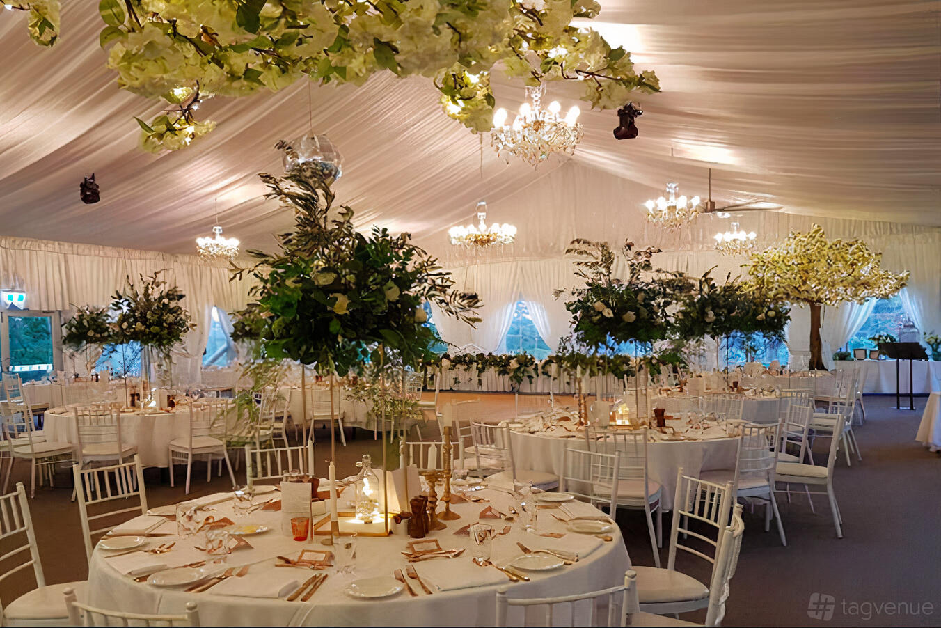 A hotel marquee with draped white ceilings, chandeliers, round tables, and tall floral centerpieces at Parklands Country Garden & Lodges.