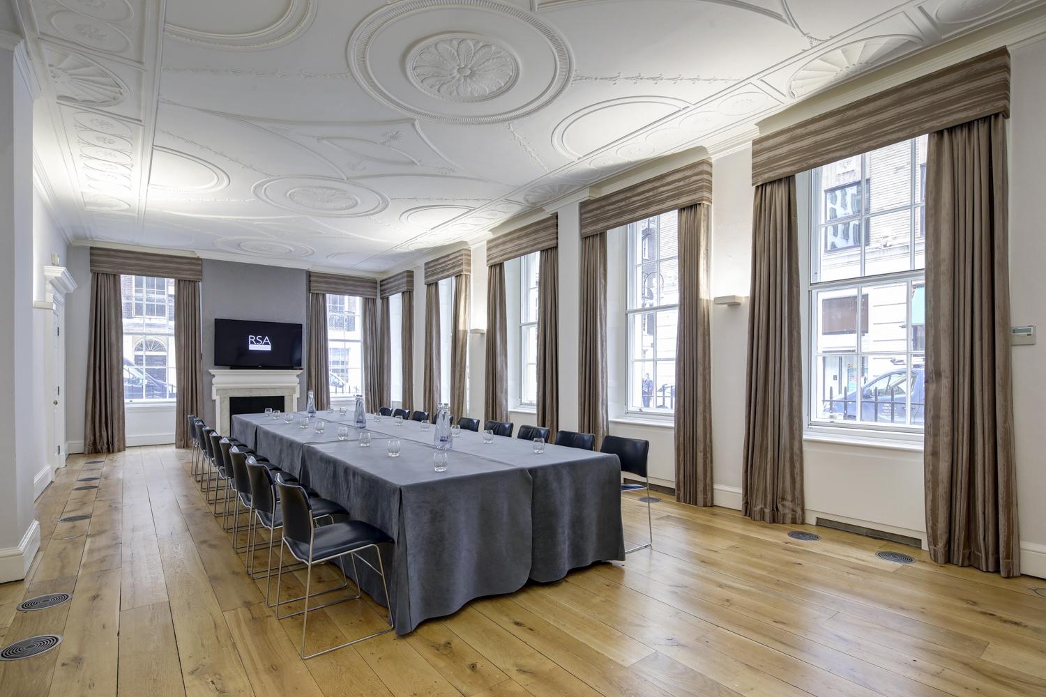 A meeting room with tall windows, hardwood floors, and a U-shaped table setup with grey linens at RSA House.