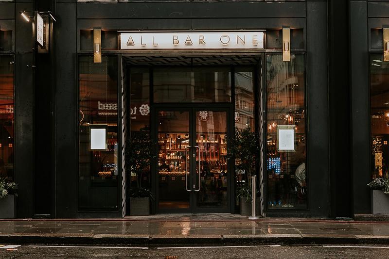 A pub entrance with glass double doors, warm lighting, and visible bar shelves at All Bar One Houndsditch.