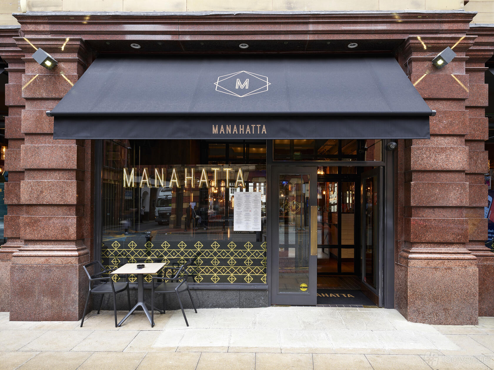 A pub with a brick facade, black awning, glass entrance, and outdoor seating at Manahatta Deansgate.