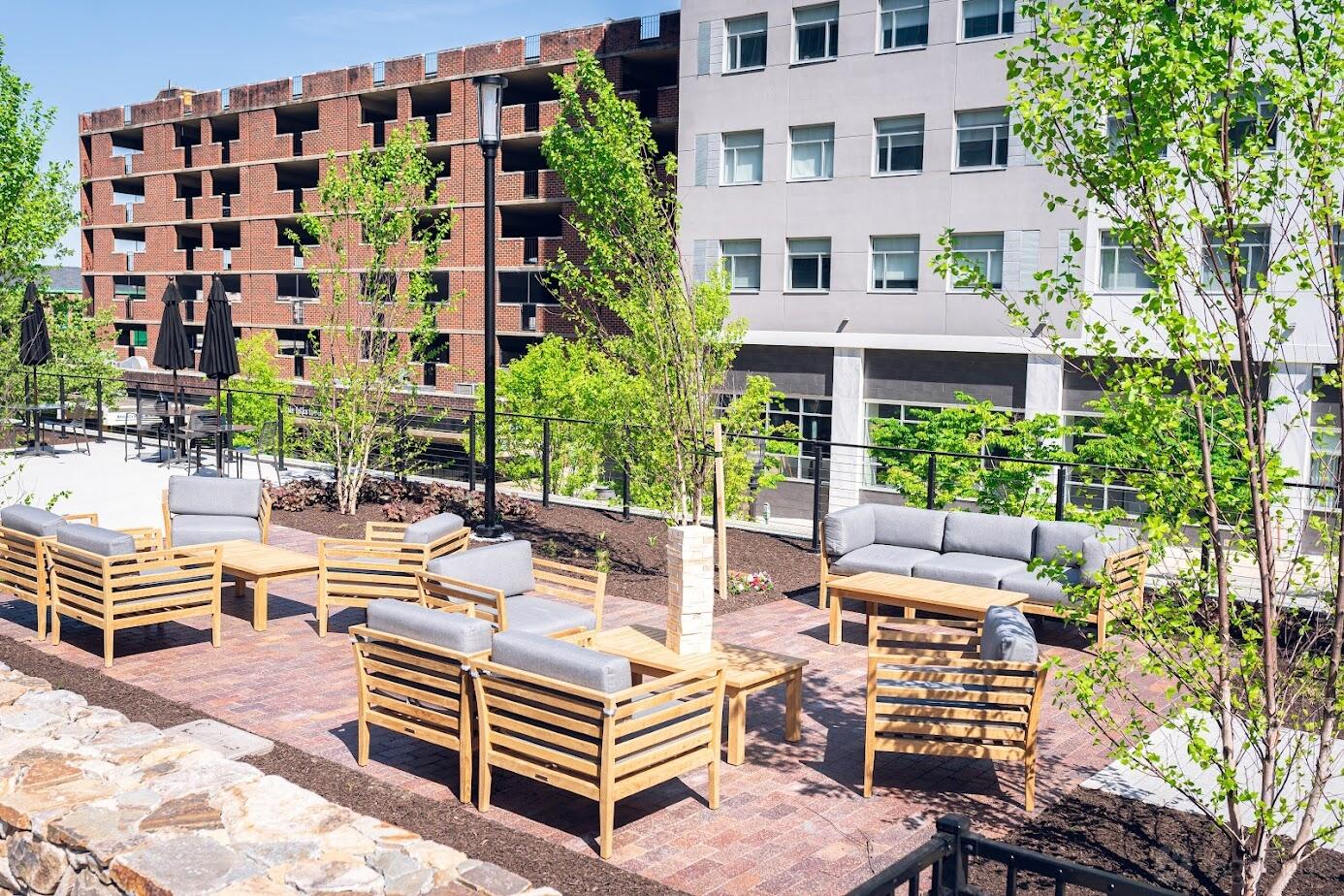 An outdoor space with wooden lounge seating, gray cushions, and young trees at The Chancery Market.