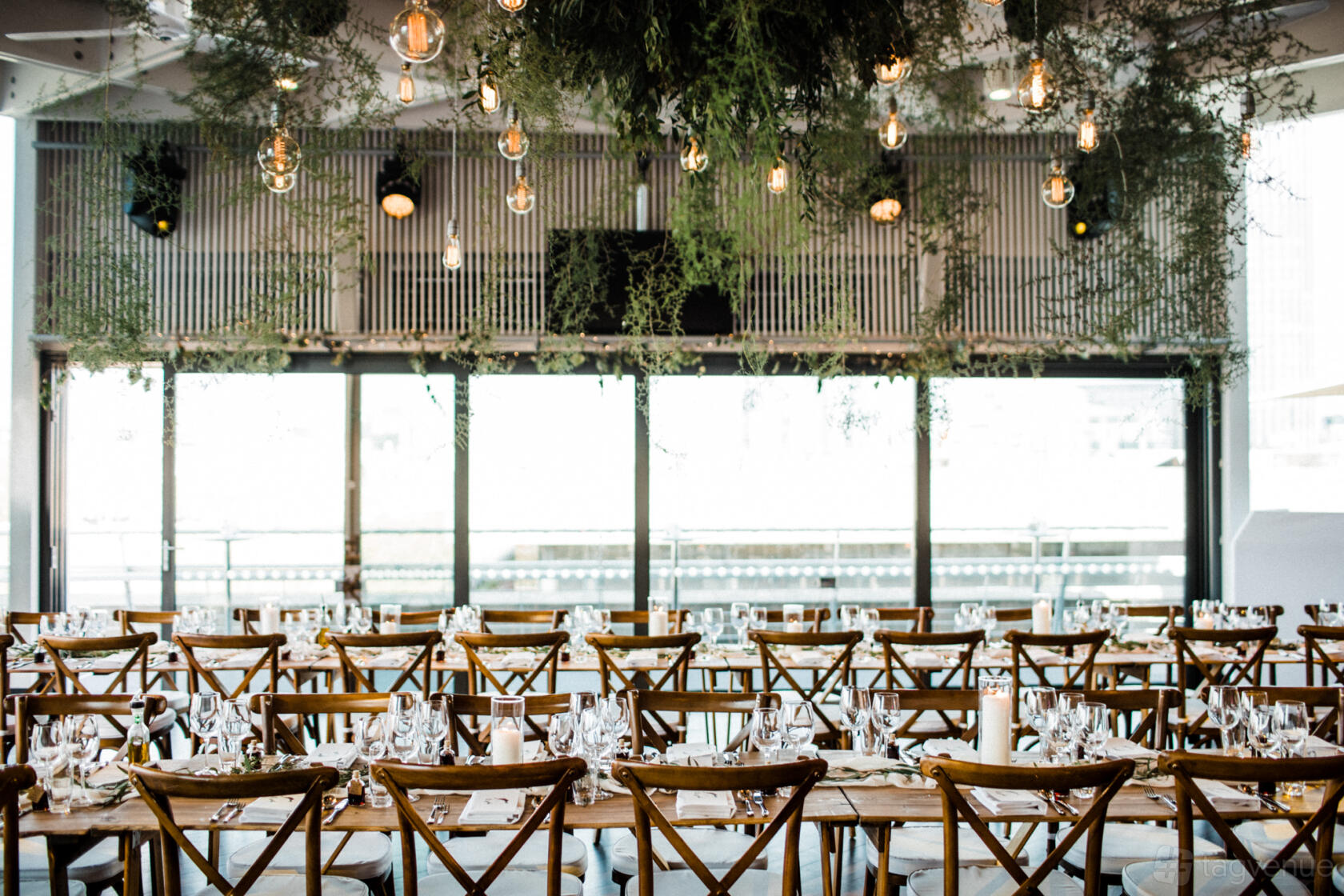 A banqueting hall with long wooden tables, cross-back chairs, hanging greenery, and glass pendant lights at The National Theatre