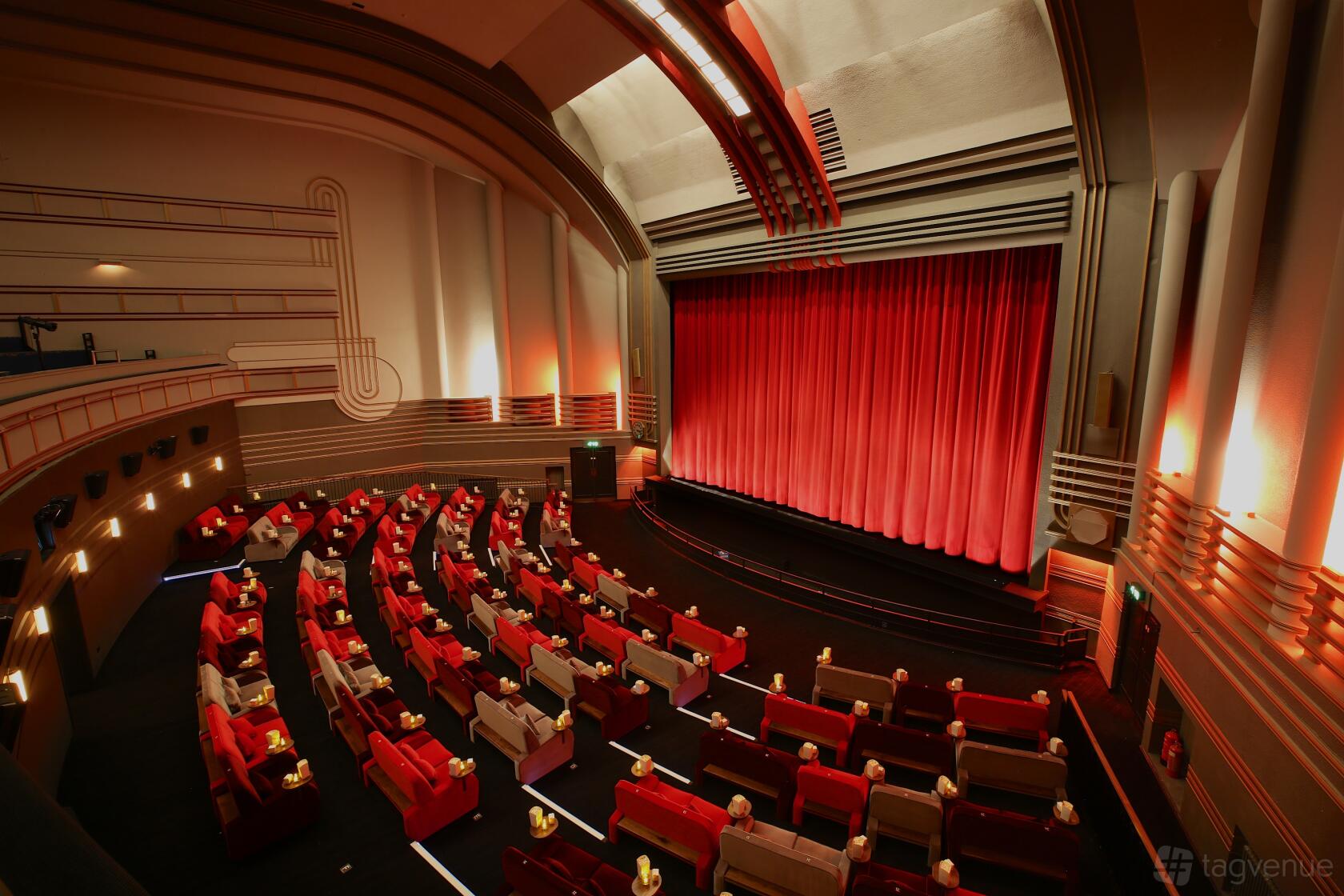 An auditorium with red plush armchairs, side tables, and a large stage with red curtains at Everyman Muswell Hill.