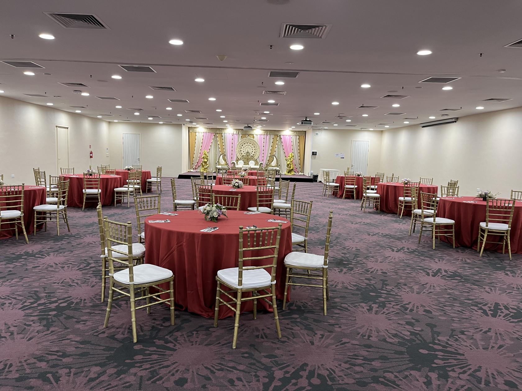 A spacious event hall with round tables draped in red linens, gold chairs, and a decorated stage at THE LEGACY, One-North.