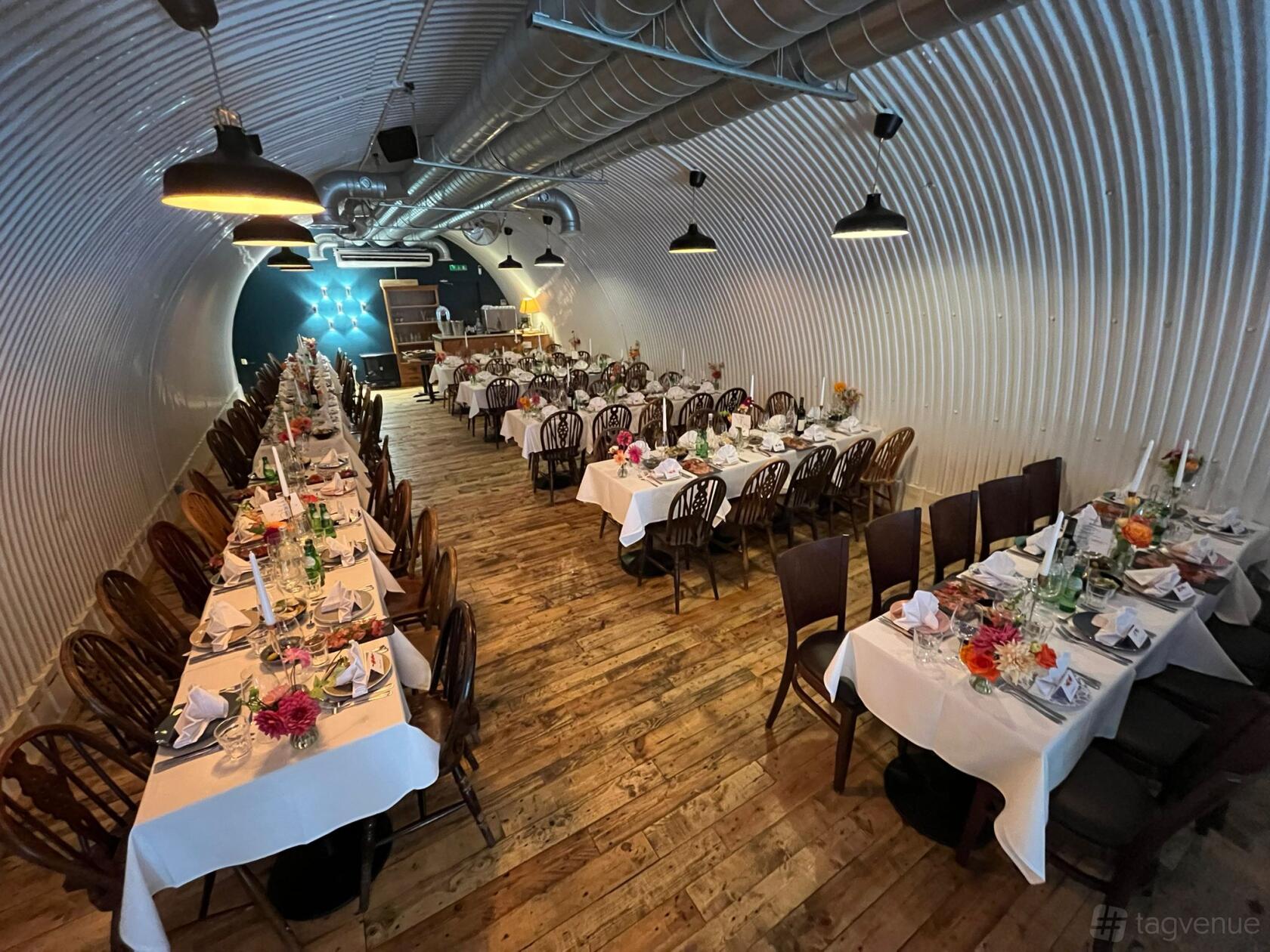 A bar with arched corrugated metal walls, exposed ductwork, and long banquet tables set for dining at Pedro's Wine Bar.