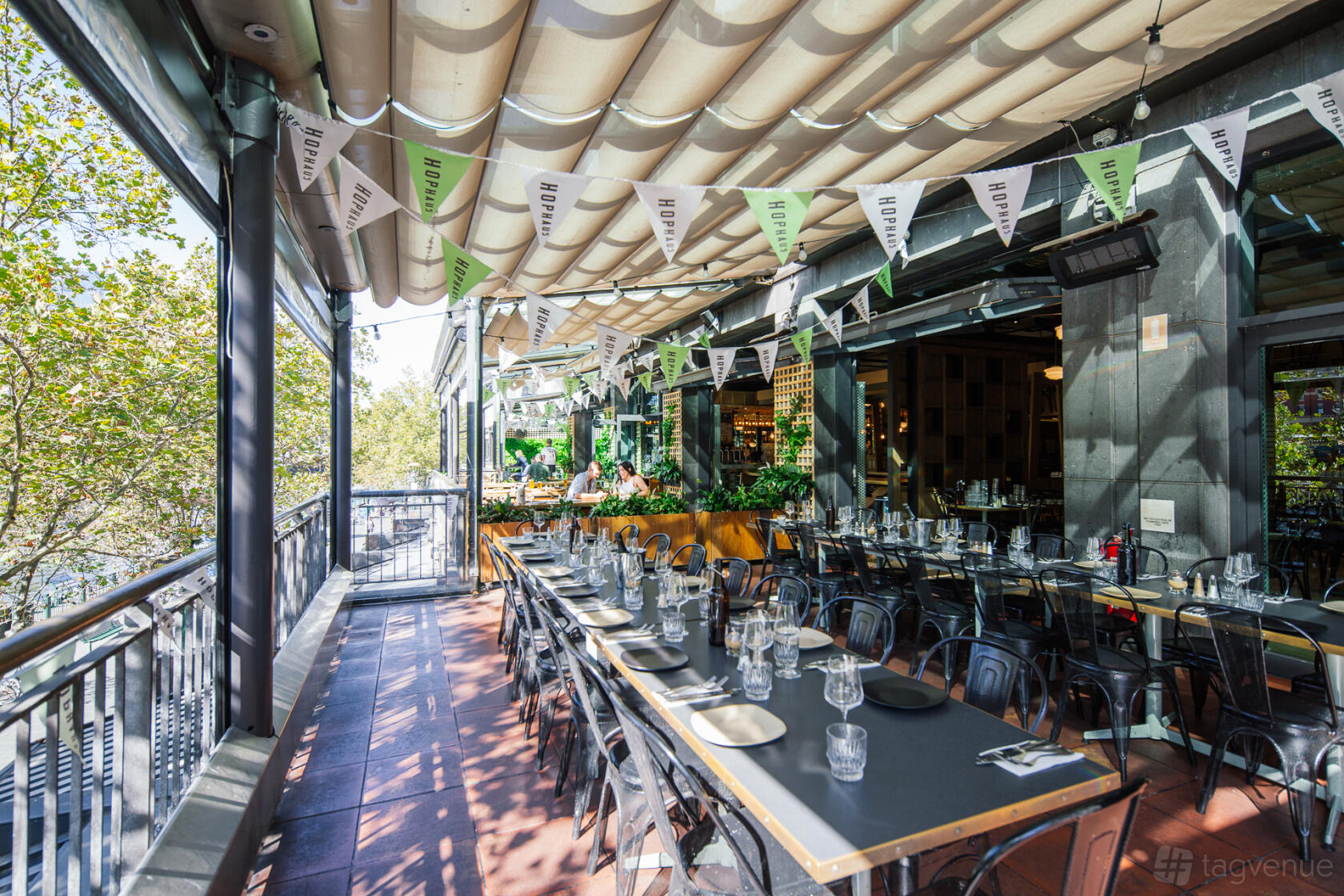 A restaurant terrace with long tables, metal chairs, and festoon bunting under a retractable roof at Hophaus.