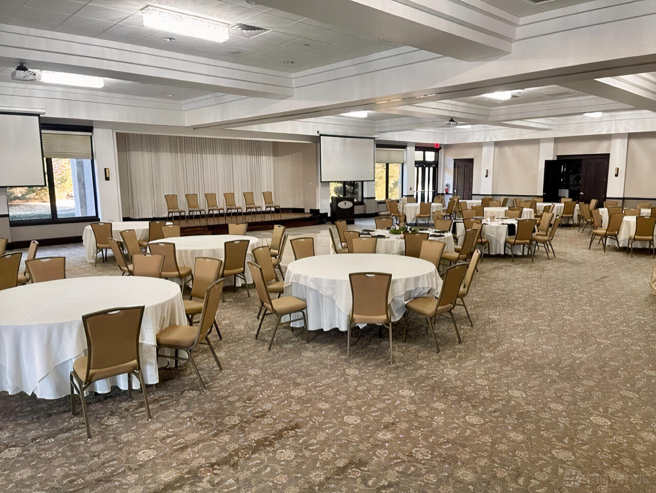 A grand ballroom with round tables, beige chairs, carpeting, and a small stage at Coyote Ridge Golf Club.