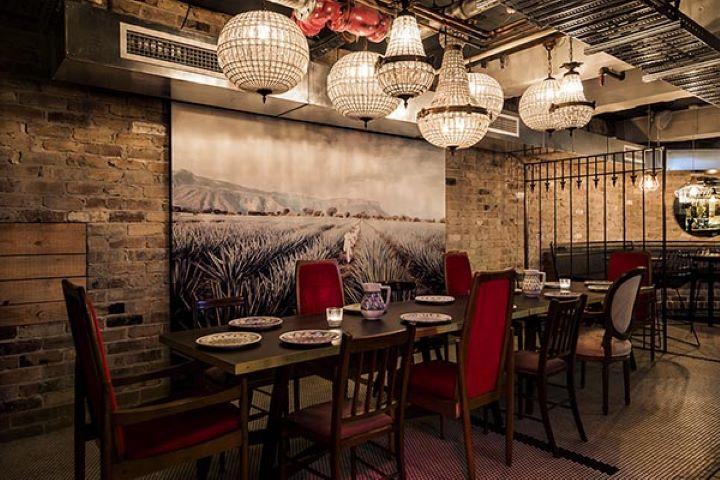A dining room with exposed brick walls, chandeliers, and a long table with red chairs at Barrio Cellar.