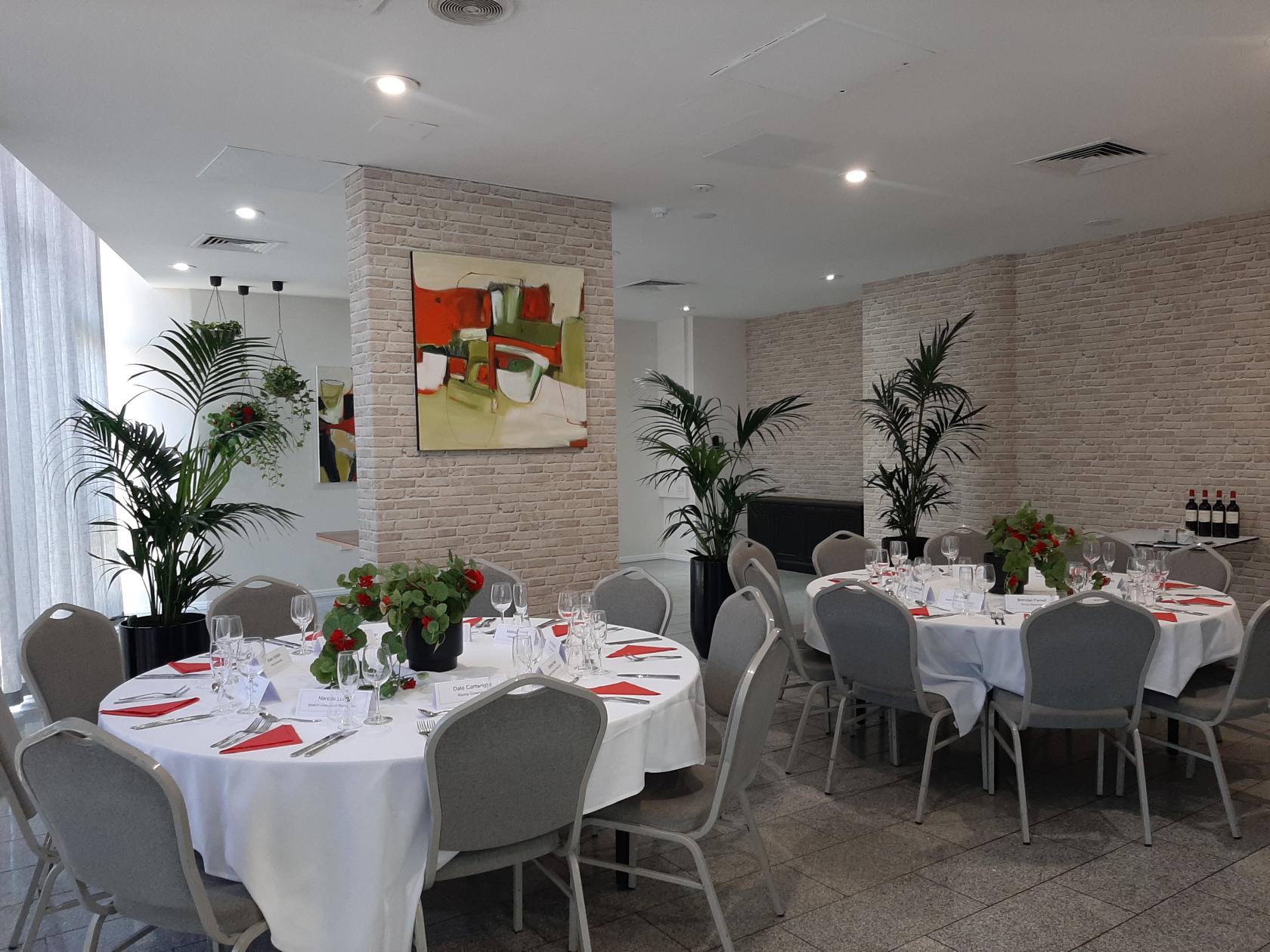 A hotel dining room with round tables set with white linens, red napkins, floral centerpieces, and wall art at Mercure Brisbane Spring Hill.