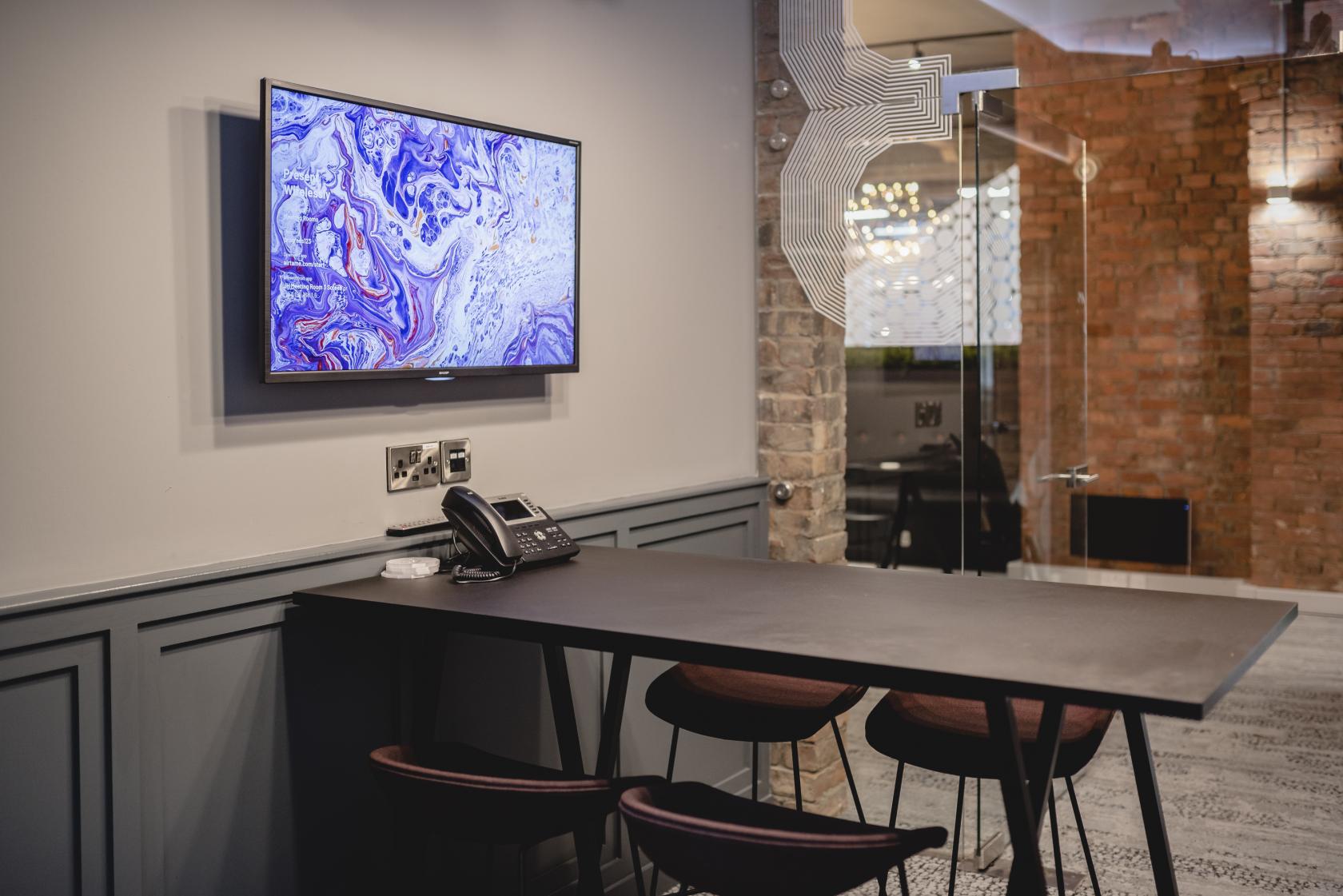 A coworking meeting room with a wall-mounted screen, bar-height table, and brick wall at Colony Jactin House.