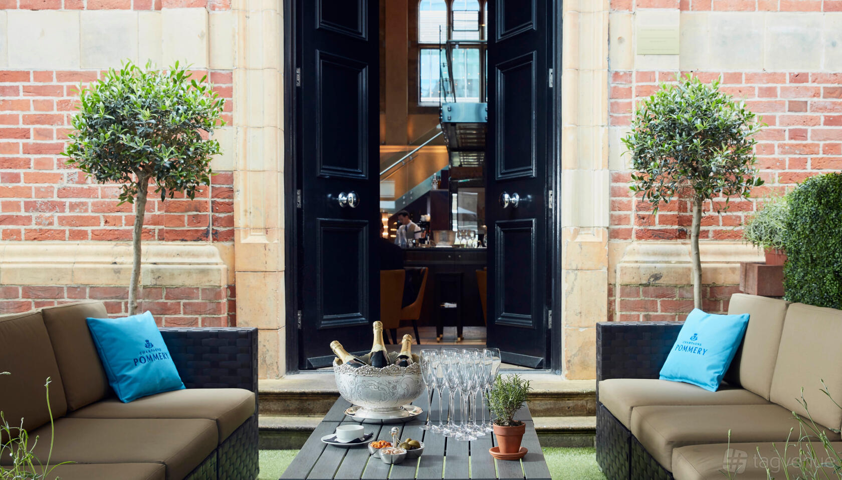An outdoor restaurant garden with cushioned seating, potted trees, and a table set with champagne and glasses at Galvin La Chapelle.