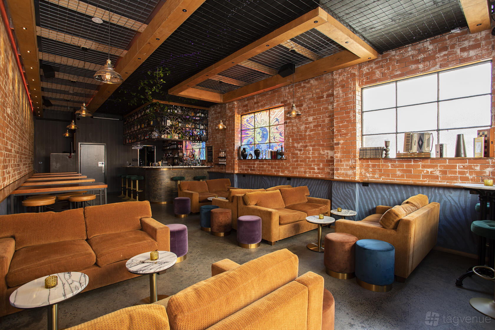 A bar with exposed brick walls, velvet lounge seating, and a stained glass window at Her.