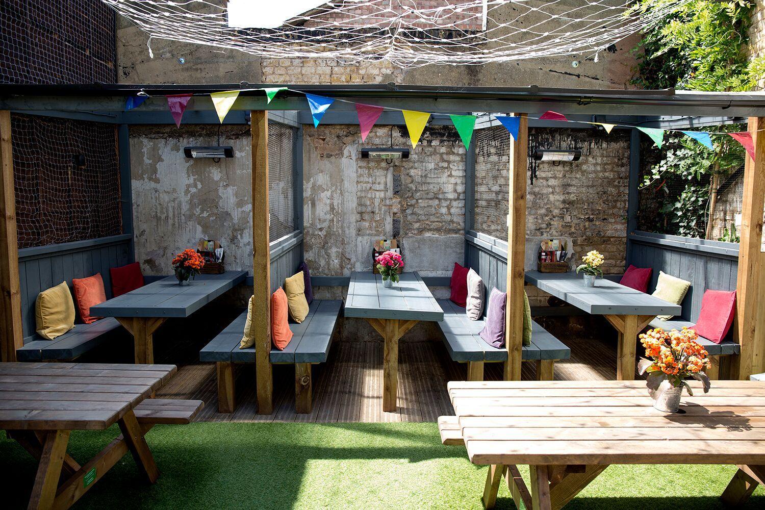 A function room in a pub with wooden picnic tables, bench seating with cushions, brick walls, and colorful bunting at The Birds.