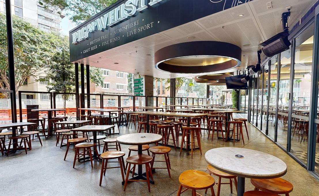 An open-air terrace restaurant with round marble tables, wooden stools, and glass walls at Pig ‘N’ Whistle - West End.