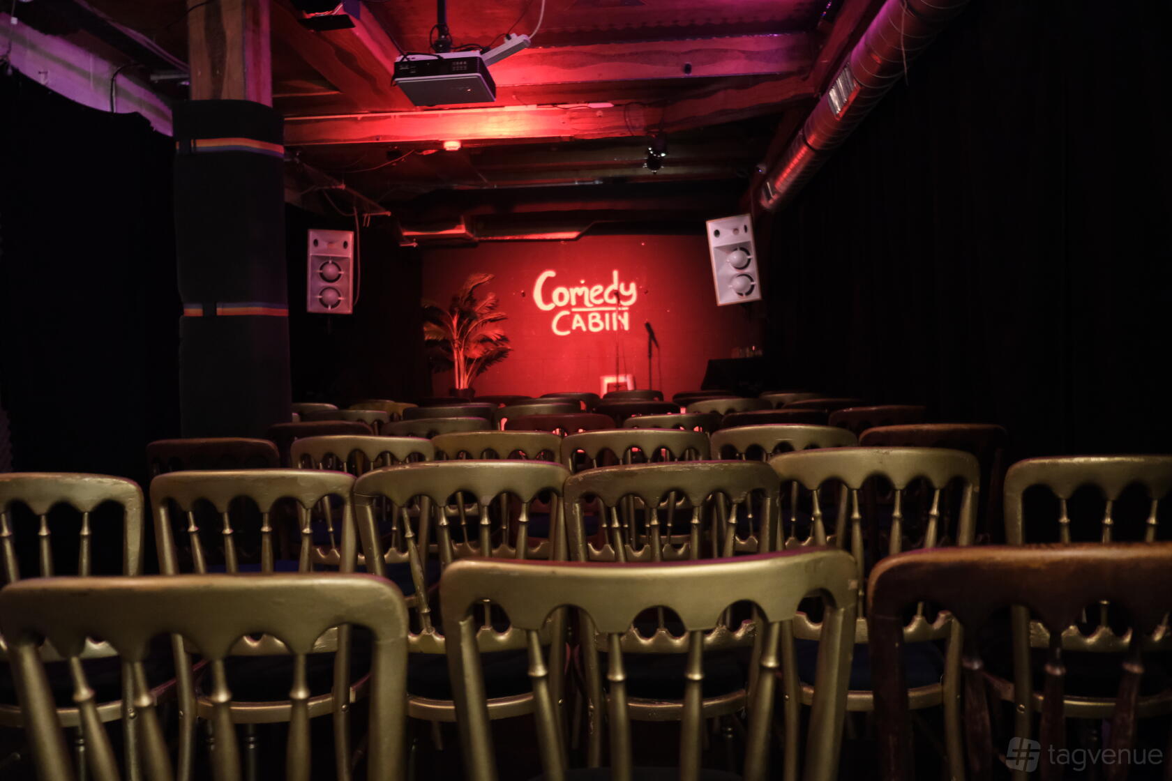 A basement event space with rows of gold chairs facing a small stage with red lighting and a neon sign at Hoxton Cabin.