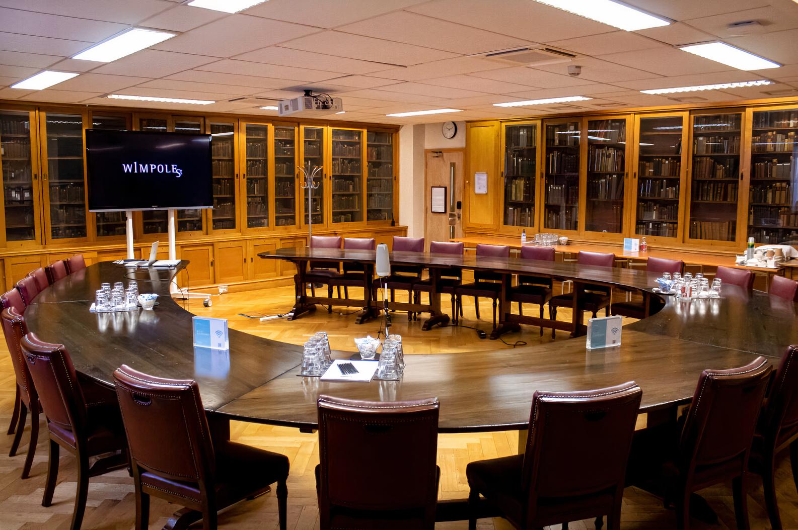 An event venue with a large U-shaped wooden table, leather chairs, and wall-to-wall bookshelves at 1 Wimpole Street.