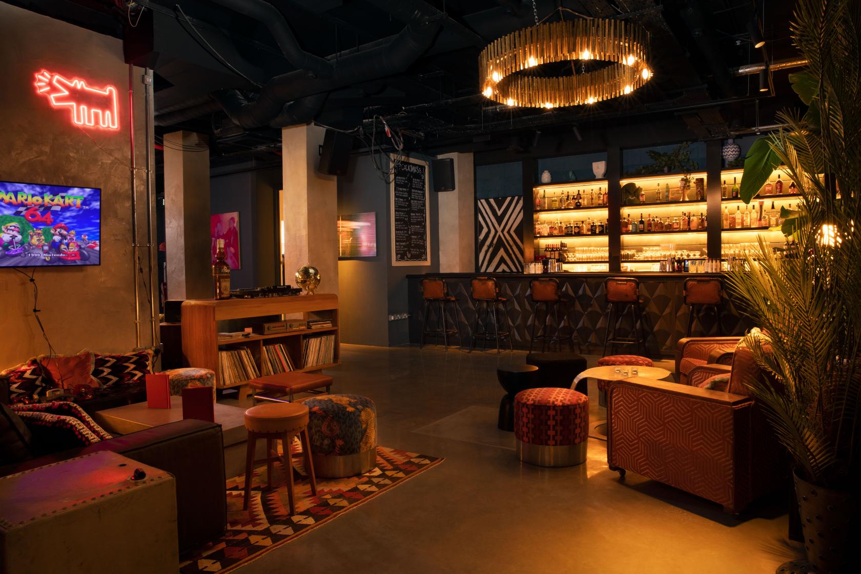 A basement cocktail bar with a backlit bar, plush seating, neon wall art, and exposed ceiling at The Little Scarlet Door.