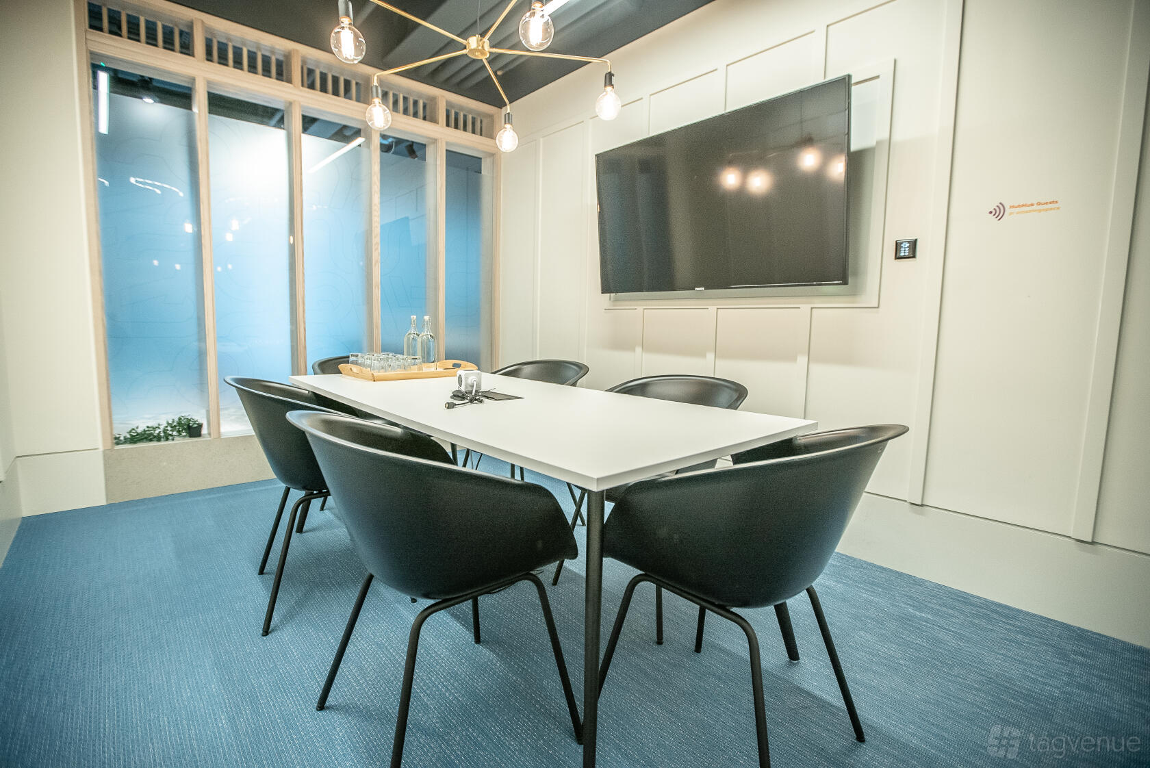 A coworking space meeting room with a rectangular table, black chairs, wall-mounted screen, and pendant lights at HubHub 20 Farringdon Street.