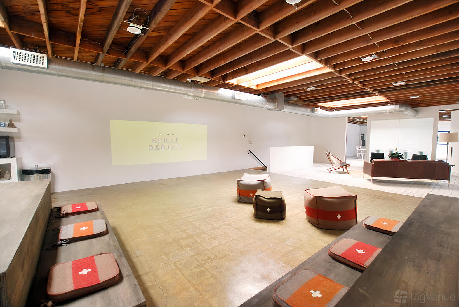 An event space with exposed wooden beams, skylights, a projector screen, and floor seating at Unita El Segundo.
