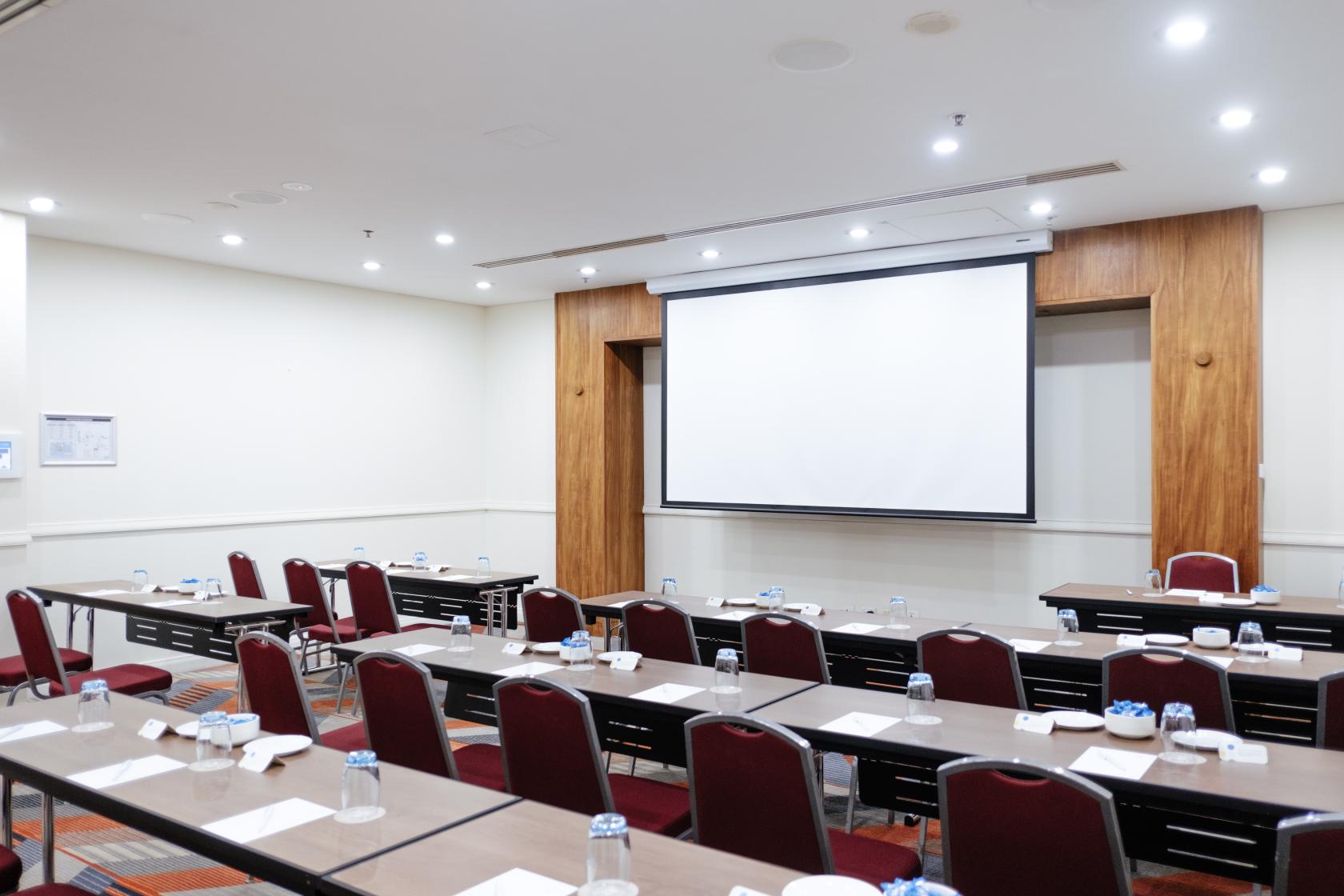 An event space with rows of tables, red chairs, a projector screen, and water glasses at Sydney Central Hotel.