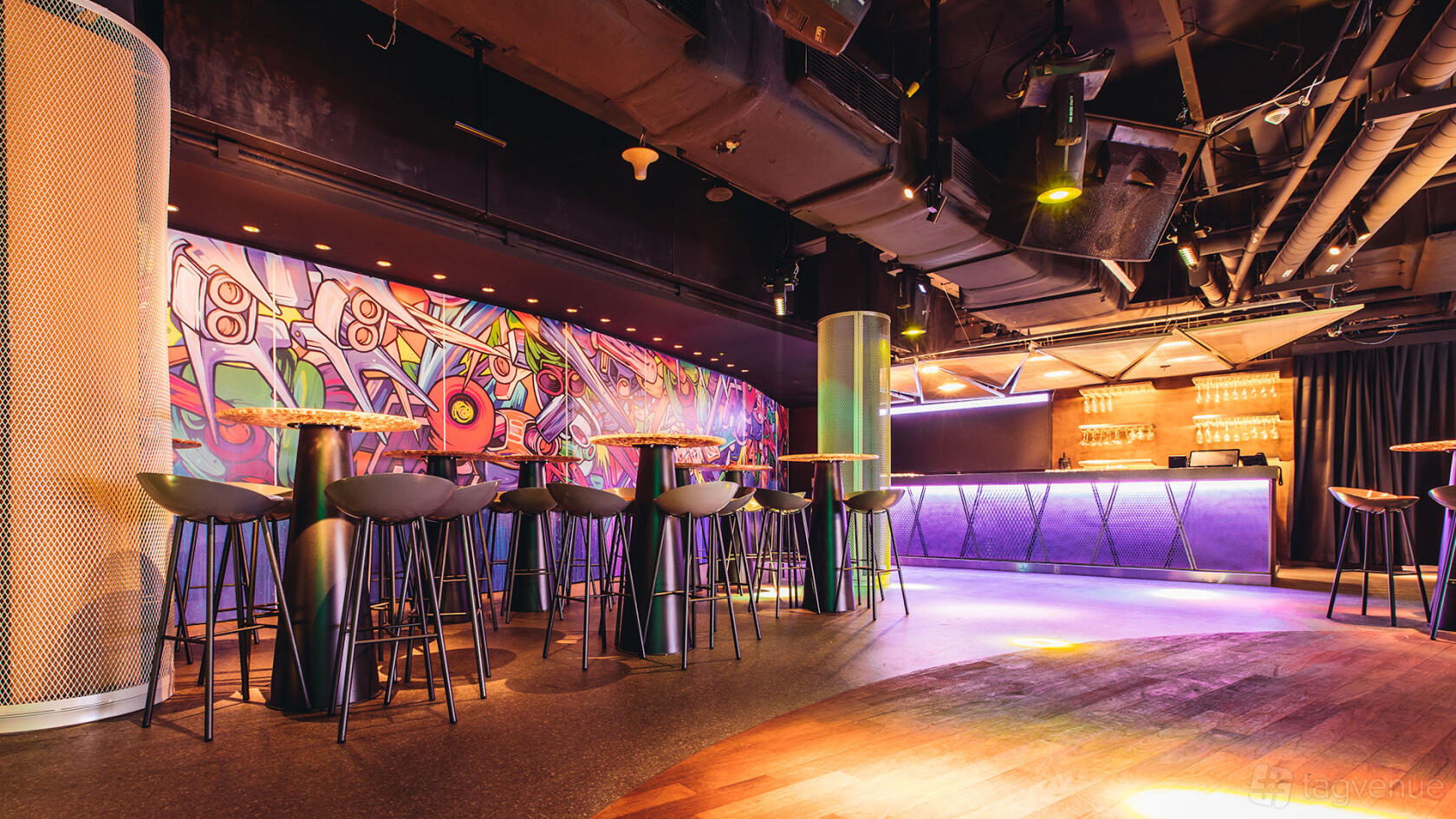 A nightclub with high tables, barstools, colorful wall mural, and illuminated bar at Phuture.