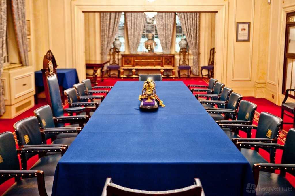 An event space in a hall with blue-covered long tables, ornate chairs, red carpet, and draped windows at Saddlers’ Hall.