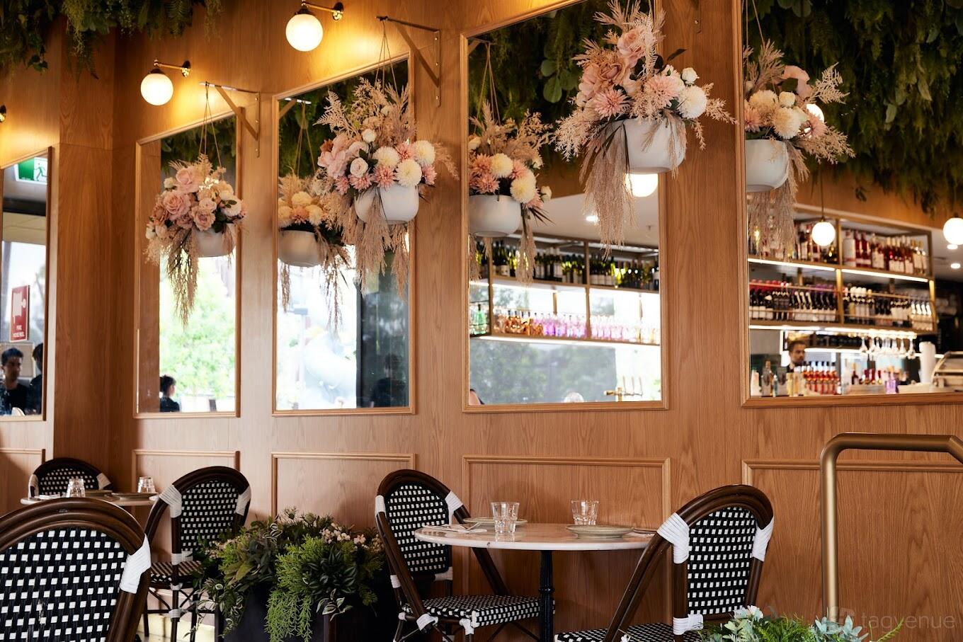 A restaurant with bistro tables, rattan chairs, wood paneling, large mirrors, and hanging floral arrangements at Garçon Lane Cove.