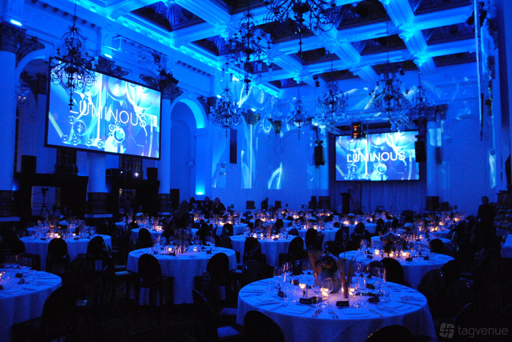 A ballroom with grand chandeliers, uplighting in blue, and round tables set for dinner at 8 Northumberland Avenue.