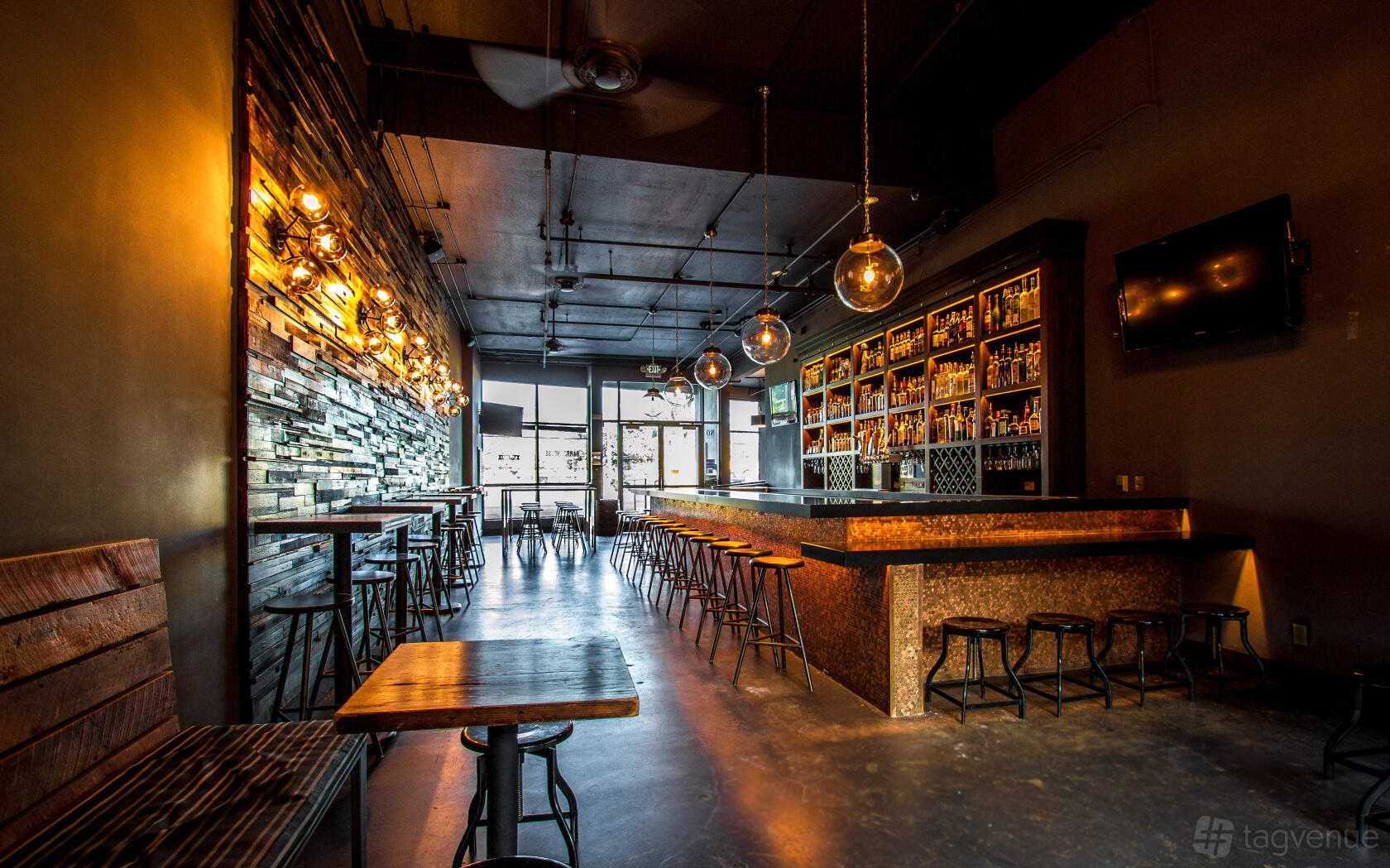 A bar with a long wooden bar counter, exposed bulb lighting, wall shelves of spirits, and wooden tables at Barrelhouse.