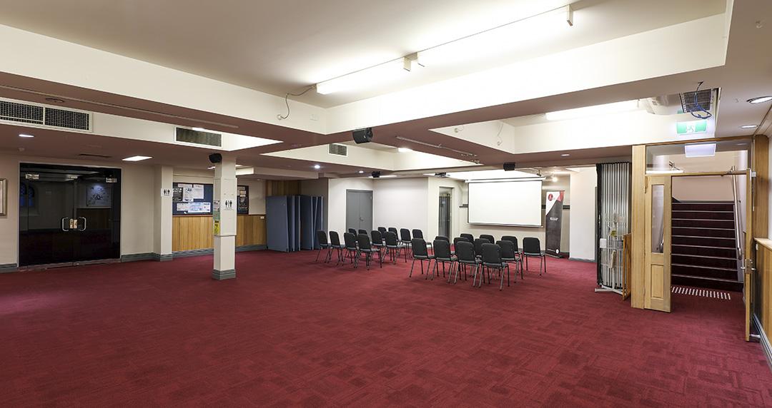 A conference hall with a red carpet, ceiling fluorescent lights, and rows of black chairs facing a projector screen at Melbourne City Conference Centre.