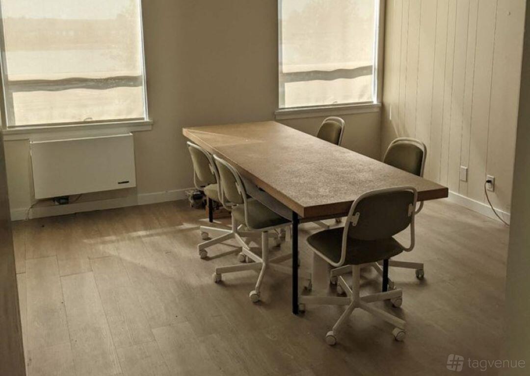 A meeting room with a rectangular wooden table, six rolling chairs, and natural light from two windows at DB Studios.