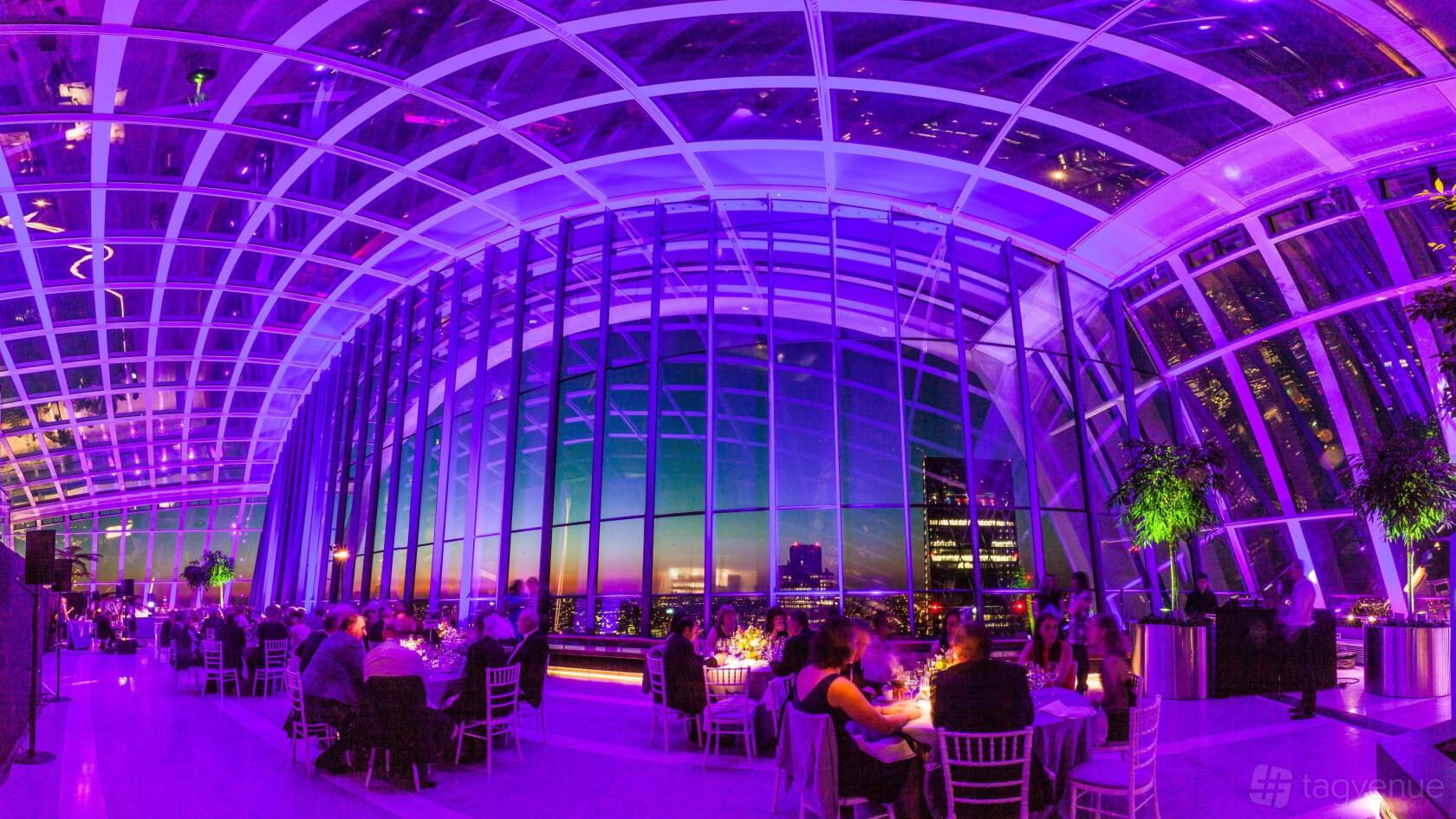 An event garden with floor-to-ceiling glass walls, purple lighting, and seated dinner tables at Sky Garden.