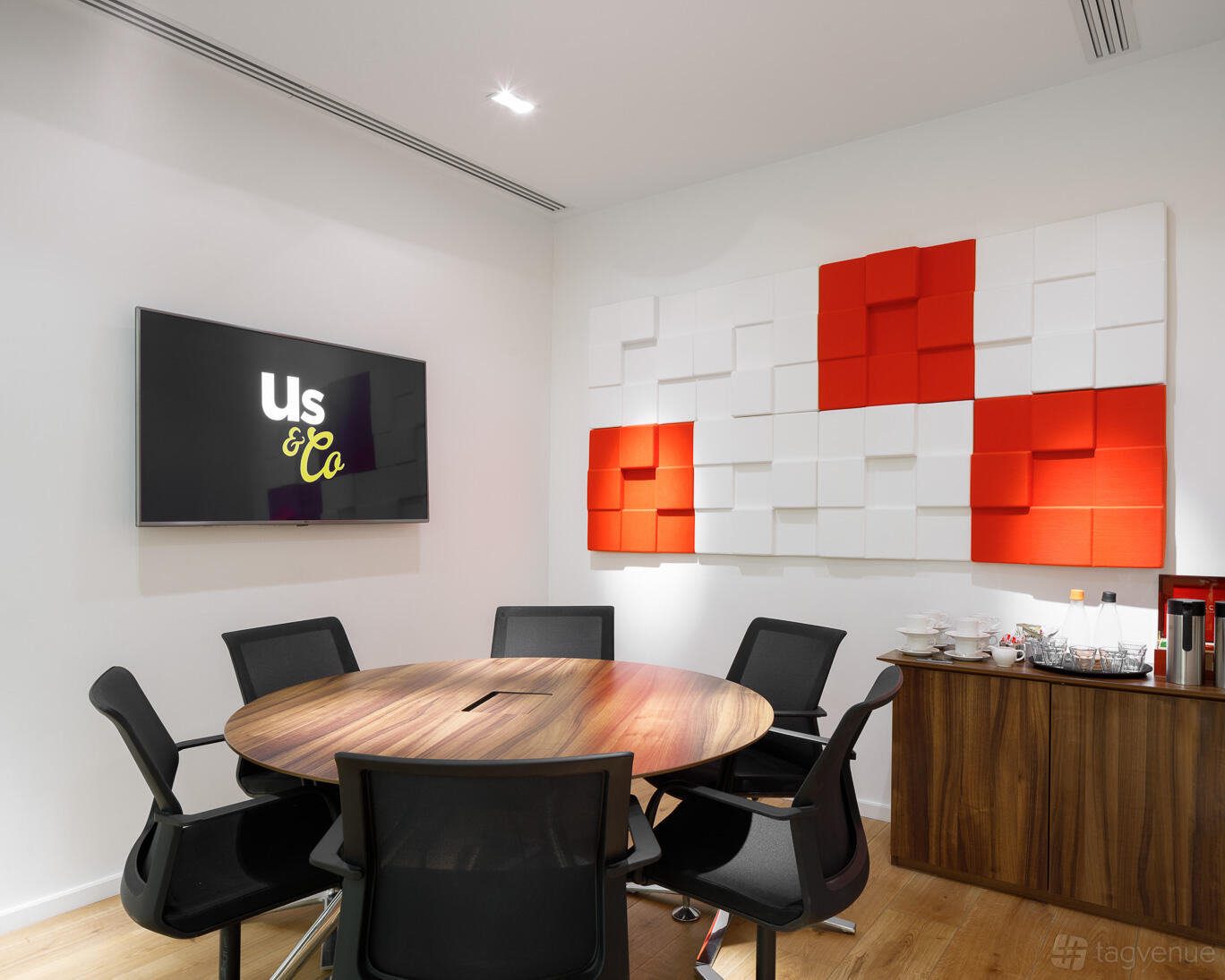 A meeting room with a round wooden table, black chairs, geometric wall panels, and a refreshment station at Us&Co Stratford.